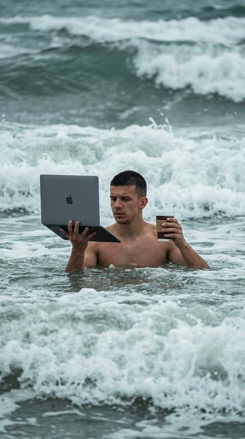 Realistic candid photo of [person from uploaded photo] standing chest-deep in rough ocean 
water, surrounded by large breaking waves and white sea foam, no hat. 
In one raised hand, held above the water, they hold an open dark grey/space-black laptop 
with a visible Apple logo, angled toward their face as if working on it. In the other hand, 
raised at chest height, they hold a to-go coffee cup with a white lid and brown sleeve.Their expression is focused and slightly tired, looking down at the laptop screen. Only the 
head, shoulders, and raised arms are visible above the waterline; the rest of the body is 
submerged. The ocean is turbulent with rolling waves and whitewater in the background, cool grey-blue-green color grading, overcast diffused daylight, slightly moody and dramatic lighting. Shot from a 
distance with a slight telephoto compression, as if photographed candidly from the shore.Vertical composition, photorealistic, ironic "always working" meme aesthetic, 
no text overlays anywhere on the image.
