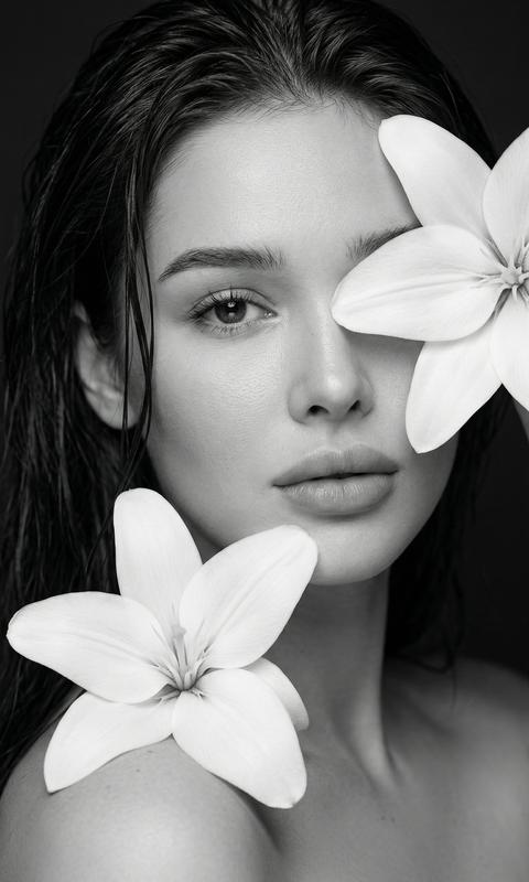 Ultra-realistic 8K fashion editorial beauty portrait. Формат 9:16; Крупный план: лицо и плечи, минимализм. Мокрые волосы, естественная текстура кожи, высокая детализация пор. Лёгкий натуральный макияж: идеальная кожа, нюдовые губы. Монохром, высокий контраст, чистый фон. Две крупные белые лилии: широкие гладкие лепестки с мягкой матовой поверхностью и выраженной формой; одна лилия частично закрывает один глаз, вторая расположена на плече, лепестки плотные, чётко очерченные, без прозрачности и деформаций. Прямой взгляд в камеру, спокойное чувственное выражение. Мягкий студийный свет, cinematic lighting, shallow depth of field, резкий фокус на глазах и коже. Профессиональная съёмка, magazine cover quality. Максимальный фотореализм, без CGI, без ретуши и анатомических искажений; лицо на 100% идентично исходному референсу — полностью узнаваемо, без искажений и без изменения пропорций, черт и внешности; любые изменения лица запрещены. Сохранить внешность
