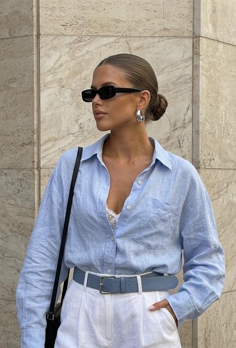 Вертикальный фотореалистичный street style fashion-портрет, средний кадр от верхней части бедер до головы: молодая взрослая женщина стоит у бежевого каменного фасада из крупных прямоугольных плит с легкой мраморной фактурой, горизонтальными швами и приглушенной городской текстурой. У нее теплый загорелый тон кожи, овальное лицо с выраженными скулами, волосы гладко зачесаны назад и собраны в низкий компактный пучок, на лице узкие черные прямоугольные солнцезащитные очки, в ушах крупные скульптурные серебристые серьги-кольца. Голова повернута влево в спокойный трехчетвертной профиль, подбородок чуть приподнят, губы расслаблены, взгляд скрыт очками; поза уверенная, сдержанная, как в уличной модной съемке. На ней свободная oversized рубашка из мягкого мятого льна бледного светлого нежно голубого  оттенка, воротник открыт, несколько верхних пуговиц расстегнуты, виден белый  кружевной бралетт или топ, на груди накладной карман, рукава длинные и объемные с расслабленными манжетами. Рубашка заправлена в белые  шорты с высокой талией, строгими складками и подвернутым низом, поверх широкий нежно голубой кожаный ремень с прямоугольной серебристой пряжкой. Одна рука в кармане шорт, другая опущена вниз рядом с тонким черным ремешком сумки у нижнего края кадра. Модель немного смещена влево, камера примерно на уровне груди с легким модным низким ракурсом, резкий фокус на лице, аксессуарах и фактуре одежды, фон остается читаемым, но вторичным. Свет естественный дневной, направленный сверху-слева, с мягкими, но заметными тенями, теплыми бликами на коже и камне; палитра приглушенная и дорогая: бежевый камень, пастельный желтый, темный коричневый, черный и серебро, реалистичный контраст, минималистичное журнальное настроение городской fashion-съемки. Без цветокоррекции фото сделано на iPhone 17 pro
