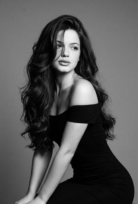 Используй лицо с моей референс\-фотографии \(face reference\), сохрани узнаваемость на 100% : не меняй овал лица , форму носа , форму бровей, губы, цвет глаз и цвет волос\. Черно‑белая студийная портретная фотография: девушка  в облегающем коротком чёрном платье без бретелек который соблазнительно подчеркивает фигуру песочные часы сидящая на табурете, повернутая в три четверти и наклонившаяся вперёд, руки опираются на колени, маникюр молочного оттенка , экстремально длинные, густые  ,очень объемные волосы в стиле супермоделей 90\-х, слоистая стрижка , укладка на брашинг с крупными мягкими волнами и сильным прикорневым объемом, глубокий косой пробор , волосы перекинуты на одну сторону , длинная прядь прикрывает часть лица , волосы блестящие с выраженным рельефом;взгляд отведён в сторону вверх ,минималистичный нейтральный серый фон; мягкое драматичное боковое освещение с едва заметным контровым волосовым светом, глубокие тени и подчёркнутые блики на коже для гламурного, соблазнительного настроения; стиль ретро fashion editorial,плёночная зернистость и сохранение текстуры волос и кожи; средний план, девушка смещена вправо по правилу третей, лёгкий низкий угол съёмки; камера портретный объектив 85 мм, диафрагма f/2\.0 — f/2\.8 для малой глубины резкости, выдержка 1/125 с, ISO 100, тонкая ретушь, чёткая детализация волос, формат 9:16