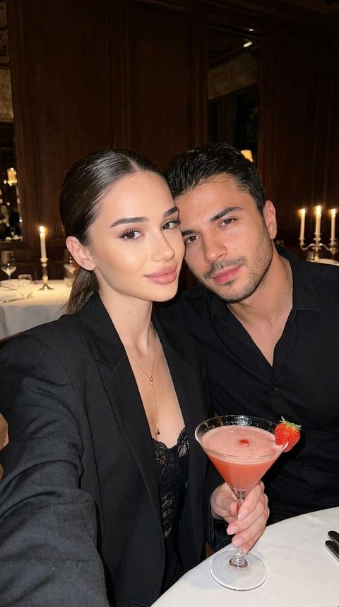 Ultra-realistic close selfie shot on a smartphone front camera (not mirror), strong direct Y2K flash, slight handheld angle, chest-up framing. A man and woman sit close together in a luxury Moscow restaurant with a dark evening atmosphere, warm candlelight and deep brown tones in the background. She wears an oversized black blazer over a black lace top with a deep neckline and a thin gold pendant necklace. Hair in a sleek low ponytail. In her hand — a martini glass with a pink cocktail and a strawberry on the rim. He wears a fitted black shirt, leaning slightly toward her. Crisp flash highlights, real skin texture, subtle grain, intimate cinematic mood, vertical 9:16.