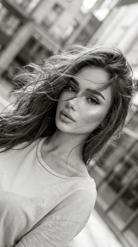 Ультра реалистичное чёрно-белое fashion video frame фото красивой девушки в движении, cinematic monochrome aesthetic, dynamic hair movement, Instagram / Pinterest editorial style, vertical frame 9:16.

Девушка снята крупным портретом под наклоном, будто кадр из видео или случайный стоп-кадр. Камера расположена близко к лицу и немного сбоку, изображение слегка повернуто по диагонали. Девушка смотрит в камеру спокойным притягательным взглядом, губы слегка приоткрыты, выражение лица уверенное, мягкое и немного загадочное.

Волосы распущенные, объёмные, живые. Волосы активно двигаются от ветра или движения головы, часть прядей летит через лицо, глаза, губы и плечи. Пряди должны выглядеть лёгкими, воздушными, немного хаотичными, с натуральной текстурой и мягким блеском. Hair in motion, cinematic movement effect.

Макияж красивый soft glam makeup: ровный сияющий тон кожи, мягкий контуринг, подчёркнутые скулы, деликатный хайлайтер на скулах, носу и над верхней губой. Выразительные глаза, длинные ресницы лисий эффект , аккуратные брови, черная  стрелка, мягкие nude shadows. Губы nude glossy lips, объёмные, с мягким контуром и сатиновым блеском. Макияж должен читаться даже в чёрно-белой обработке.

На девушке светлый футболка, мягкая ткань, минималистичный городской образ. Верх одежды слегка открыт у шеи и ключиц, образ элегантный, не перегруженный.

Свет мягкий дневной, natural city daylight, без вспышки. Свет идёт сбоку и сверху, подсвечивает лицо, волосы, губы и ключицы. На волосах светлые highlights, на коже мягкое сияние. Тени плавные, без жёсткой студийности. Свет выглядит как естественная съёмка на телефон на улице или возле городской архитектуры.

Цветокоррекция чёрно-белая, soft monochrome grading, мягкие серые полутона, светлые highlights на волосах, глубокие, но не проваленные тени, low contrast editorial look, slight blur from movement, subtle film grain. Кадр должен выглядеть живым, немного размытым, как стильный стоп-кадр из Reels.

Фон городской, сильно размытый: архитектура, улица, фасады зданий, стекло или витрины. Background blur, shallow depth of field, город не должен отвлекать, главный акцент на лице, волосах и движении.

Композиция: close-up / medium close-up shot, diagonal tilted frame, лицо девушки занимает центр кадра, волосы пересекают кадр динамичными линиями. Вертикальный формат 9:16, ощущение движения, лёгкая неидеальная резкость, aesthetic mobile video frame.

Качество: снято на iPhone 17 Pro, ultra realistic mobile photography, cinematic black and white video frame, realistic skin texture, detailed moving hair strands, natural motion blur, soft focus, 4K, без мультяшности, без plastic skin.

Negative prompt:
color photo, perfectly still hair, slick bun, harsh flash, studio background, looking away, wide smile, teeth showing, overexposed face, messy low quality blur, plastic skin, cartoon face, CGI look, distorted eyes, distorted face, bad anatomy, extra fingers, low quality.