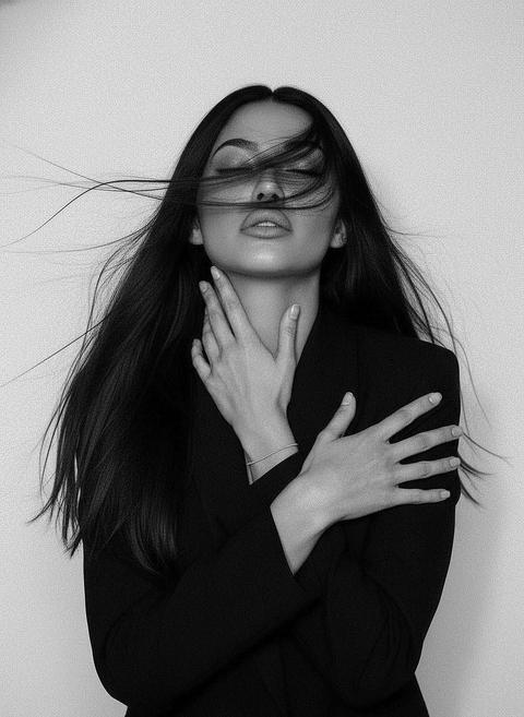 Reference image: conceptual variant.
Black and white artistic portrait, medium-close framing. Девушка занимает центр кадра, корпус слегка развернут, плечи видны. Голова наклонена назад и немного в сторону, подбородок вытянут, глаза закрыты, выражение лица спокойное и глубокое, с намёком на внутреннее напряжение. Hair: long, loose, wind-swept strands crossing the face and cheekbones, несколько прядей плавно тянутся по воздуху. Pose: обе руки подняты к шее и ключицам. Одна рука касается шеи, пальцы вытянуты, вены и анатомия видны. Вторая рука пересекает грудь, ладонь лежит на плече, пальцы длинные, естественные, ногти короткие без акцента. Запястье с тонкой едва заметной нитевой резинкой. Outfit: structured black blazer with sharp minimal silhouette, без видимых деталей, ткань matte, контрастная к коже. Фигура читается через форму и негативное пространство. Lighting: high-contrast soft directional light, smooth gradients, sculpted cheekbones, deep shadows under jawline. Background: clean white, seamless, no texture. Camera: medium close-up, straight-on angle, shallow depth of field, crisp detail on face and hands, slight grain for analog feel. Mood: raw elegance, introspection, tension, wind-driven stillness, fashion editorial minimalism.