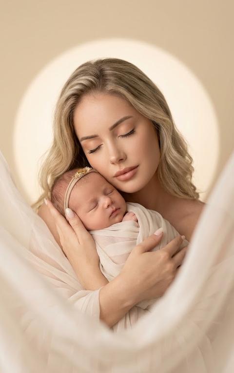 A woman holds a sleeping newborn to her chest, her eyes closed and her head slightly tilted toward the baby. The woman is turned to the camera at a three-quarter angle. A close-up portrait from chest to head, shot at eye level, the composition is centered, mother and child completely fill the frame, with translucent flowing fabric smoothly surrounding them, creating soft, airy lines around the composition. The main focus is on the faces of the mother and baby. Focus priority: faces.

The skin looks perfectly even and matte. Light beige-peach shadow on the eyelids, thin brown eyeliner along the lash line, and long natural lashes with mascara. The lips are covered with a warm nude satin lipstick (RGB #C68F79) without a pronounced shine.

The woman's hair is styled in large waves and tossed to one side.

The manicure is medium-length, soft oval shape, translucent milky finish (RGB #F7F2EE) with a glossy finish. The mother and child are completely enveloped in a long, translucent, lightweight, milky-colored chiffon with a soft, matte finish. The fabric drapes loosely around the shoulders, arms, and child, creating soft, airy folds. The baby's head is adorned with a thin, elastic headband with a small, gold-toned decorative element (RGB #D4AF37).

The shoot takes place in a minimalist studio with a warm, creamy-beige background (RGB #E9DDC5). A soft, barely-there circle of light is positioned behind the model, creating a delicate glow around the silhouettes of the mother and child. The translucent fabric flows in front of the lens and around the models, creating a sense of lightness and volume without obscuring their faces.

The studio lighting is based on a single, large, soft light source positioned slightly above eye level and slightly off to the side. The light evenly illuminates the faces of the mother and child, highlighting the soft transitions of skin and the volume of the fabric. Additional reflected light gently fills shadow areas. The shadows are very soft and smooth. The contrast is low. The luminous background creates a subtle halo around the silhouettes, while the chiffon takes on a translucent, airy texture with delicate light transitions.

Shot with an 85mm portrait lens. Shallow depth of field. Focus on faces. Maximum sharpness is concentrated on the faces of the mother and newborn, along with skin texture, eyelashes, and folds of fabric. The foreground and background are softly blurred, creating a creamy bokeh. Fine art maternity portrait photography, newborn photography, highly detailed, photorealistic.