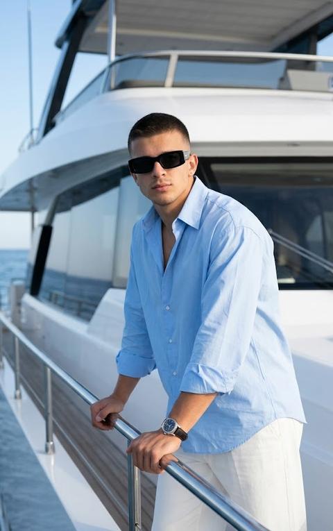 A man stands on the deck of a modern luxury yacht, leaning slightly forward and resting both hands on the polished metal handrail. His body is slightly turned toward the camera, his head is raised, and his gaze is directed directly at the lens through sunglasses. The shot is taken from the hips to the head, at eye level, the composition is centered, and the architectural lines of the yacht create a minimalist background and accentuate the model's figure. The main focus is on the man's face. Focus priority: focus on the face.

The man is wearing a loose, oversized shirt made of smooth cotton poplin with a slight satin sheen in a sky-blue shade (RGB #C8DCF2). The collar is unbuttoned, and the sleeves are casually rolled up to the forearms. The lower half of the look consists of wide-leg trousers made of thick cotton twill with a matte finish in a white shade (RGB #F5F4EF). He is wearing rectangular sunglasses with wide black acetate frames (RGB #1F1F1F) and dark lenses with subtle mirrored reflections. On her wrist is a watch with a metal case and a dark leather strap.

The shoot takes place on the deck of a modern motor yacht with a minimalist design. The frame features the crisp white glossy surfaces of the hull, polished stainless steel handrails, large panoramic windows, and the vessel's smooth architectural lines. A clear, cloudless blue sky can be seen in the background, emphasizing the atmosphere of a premium seaside vacation.

The lighting is natural, bright daylight. The main light source is positioned above and to the right of the model, creating a distinct, directional light. The shadows are dense and graphic, the contrast is high, yet the details in the highlights of her clothing and the yacht's hull are fully preserved. The metal surfaces and glass create controlled, bright highlights, and her skin takes on a warm, natural tone.

Shot with an 85mm portrait lens. Shallow depth of field. Focus on the face. The man's face is sharply defined, with the textures of the cotton fabric, metal handrails, eyeglass frames, and leather well rendered, while the yacht's architectural elements gradually soften thanks to a shallow depth of field. Luxury lifestyle photography, fashion editorial style, highly detailed, photorealistic.

Clean luxury color grading, bright natural sunlight, warm neutral tones, crisp whites, deep blue sky, moderate saturation, high contrast, sharp directional light, clean shadows, glossy highlight roll-off, premium editorial aesthetics, cinematic luxury look, high clarity, realistic skin tones, subtle color separation, modern coastal lifestyle, highly detailed, photorealistic, polished commercial finish