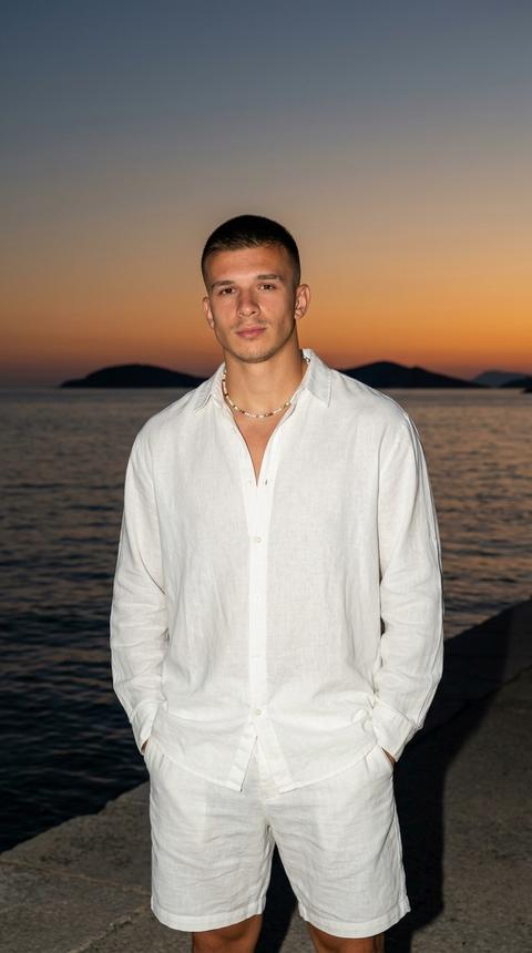 A man stands on the embankment at the water's edge during sunset, his shoulders relaxed, his hands in his pockets. He looks directly into the camera. The shot is taken from mid-thigh to head, at eye level, the composition is centered, the figure occupies the center of the frame, and the horizon and sunset sky form the background. The main focus of the shot is the man's face, his white shirt, and the contrast between the illuminated figure and the evening seascape.

The man is wearing an oversized pure white (#F8F8F6) linen shirt. The top buttons are undone, the collar is open, and the fabric forms natural folds along the torso and sleeves. The lower part of the image—wide bras—is made in the same light color scheme and fabric. The clothing appears light and appropriate for warm weather by the sea. Around his neck is a thin necklace of multicolored beads.

The scene takes place on the seaside promenade in the evening. In the background is a calm water surface with gentle waves. The dark silhouettes of islands and rocky outcrops are visible on the horizon. The sky is painted in a smooth gradient from deep blue at the top to a rich orange-coral hue (#E97A4D) near the horizon. The coastline and distant objects remain minimalist, not distracting from the model.

The lighting is based on a combination of natural sunset light and a soft front-facing flash. The flash is positioned close to the camera and evenly illuminates the man's face, neck, and shirt, preserving the subject's high detail. The sunset sky and sea background remain slightly darker than the foreground. Shadows on the face are soft and shallow. The contrast between the illuminated subject and the surrounding environment is moderately high. The highlights on the shirt are clean and controlled, without blown-out highlights.

Shot with an 85mm portrait lens. Shallow depth of field. The focus is on the man's face and the top of his shirt. The sea background and horizon line remain slightly blurred, preserving the readability of the surrounding environment. High detail of skin, shirt fabric, water surface and sunset sky. Coastal editorial portrait photography, highly detailed, photorealistic.

cinematic sunset coastal mood, warm orange and deep blue dominant tones, moderate saturation, medium-high contrast, flash-lit subject with darker ambient background, clean highlights, rich sunset gradient sky, soft shadow transitions on skin, natural skin tone rendering, crisp white clothing separation, smooth water reflections, minimal grain, editorial evening color grading, polished luxury vacation photography look.