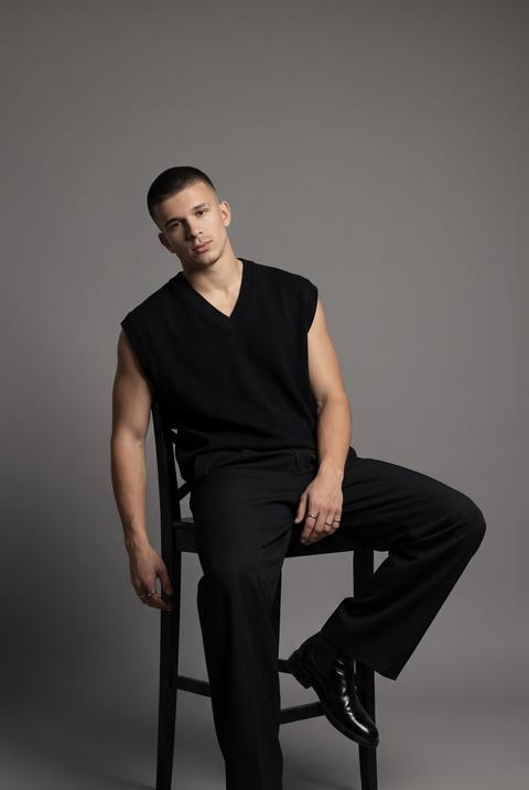 A photograph of a man seated on a tall black wooden chair in a studio, his body turned three-quarters, one leg extended forward, the other bent and extended to the side, one arm hanging loosely by his side. His head is slightly tilted, looking directly at the lens. The shot extends from head to toe, with the camera positioned at eye level.

The man is wearing a black, oversized, sleeveless vest made of thick, smooth fabric with a matte finish and a V-neck. He is wearing black, loose-fitting, wide-leg trousers made of thick, matte fabric with soft, natural folds at the hips and knees. He is wearing black, leather boots with a smooth, glossy finish and a rounded toe.

On his fingers, he wears several thin, silver-tone metal rings. The shoot takes place in a minimalist studio with a monochromatic gray-graphite background (RGB 75, 75, 75). The wall and floor are visually united into a single space with no discernible horizon line.

A professional soft studio light is used. The light is directed at an angle onto the face and upper body, creating a clear three-dimensional modeling of the face and hands. The left side of the face and body is brightly lit, while the right side gradually transitions into soft shadow. The shadows are medium density with soft edges.

Shot with an 85mm lens, medium depth of field, focusing on the eyes and face. The face and upper body are extremely sharp and detailed. A realistic portrait perspective is used, highly detailed, photorealistic, cinematic editorial fashion photography.