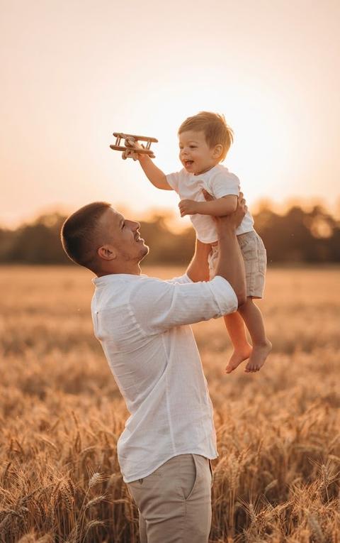 > Трогательный семейный портрет в стиле «father and son bonding» на фоне пшеничного поля. Мужчина стоит в профиль, держа маленького ребенка на руках высоко над собой. Мужчина смотрит на ребенка с любовью и легкой улыбкой. Ребенок сидит на руках, протянув одну руку вверх и держа деревянную игрушечную модель самолета. Лицо ребенка обращено к игрушке, выражение радости и восторга.

> Одежда: Образы в единой светлой, натуральной гамме. На мужчине — простая белая футболка и светло-бежевые брюки чинос. На ребенке — белая футболка и светлые бежевые шорты.

> Окружение и освещение: Локация — высокое поле спелой пшеницы. На заднем плане размытая линия горизонта и силуэты деревьев. Освещение — яркий, теплый свет «золотого часа» (закатное солнце). Солнце находится за спинами моделей, создавая мощный контровой свет и золотистый ореол (rim light) на контурах фигур и колосьях пшеницы. Атмосфера теплая, летняя, полная нежности.

> Технические параметры: Съемка на профессиональную камеру с портретным объективом 85mm. Средний план в полный рост. Открытая диафрагма f/1.8–f/2.0 для создания глубокого эффекта боке (художественного размытия поля на заднем плане) при сохранении идеальной резкости на фигурах. Теплая кинематографичная цветокоррекция, высокая детализация, стиль family lifestyle photography, формат 8k, фотореализм.
