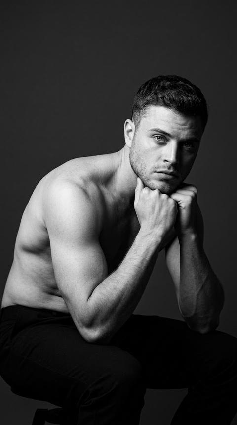 Black and white portrait. The man sits sideways to the camera, leaning slightly forward. His hands are raised and under his chin, looking directly into the lens. The shot is a close-up portrait from waist to head, with the camera positioned at eye level. The composition is minimalist, with plenty of negative space, highlighting the body's shape and the play of light. The main focus is on the face, eyes, and hand. Focus priority: face.

The natural contours of the face, shoulders, and arms are clearly visible, while maintaining realistic skin texture.

The upper body is completely bare, showcasing the natural anatomy of the shoulders, chest, and arms. The model wears dark trousers made of a dense matte fabric in a deep black shade (RGB #171717), which practically dissolve into the shadows of the frame.

The shoot was taken in a professional photo studio against a monochromatic dark gray background (RGB #2A2A2A) with a completely minimalist environment without any decorative elements. All attention is focused solely on the model and the light and shadow pattern.

The studio lighting is based on a single, hard, directional light source positioned from the top left of the model at an angle of approximately 45 degrees. The light creates expressive volumetric modeling of the face and body, highlighting the muscles of the arms, shoulders, and collarbones. The shadows are deep and clearly defined, with high contrast. The highlights remain clean, without any flare. The right side of the face and body gradually fades into dense shadow, enhancing the drama of the image.

Shot with an 85mm portrait lens. Shallow depth of field. Focus on the face. Maximum sharpness is concentrated on the eyes, facial skin, hands, and the texture of the shoulders, while the background remains soft and completely uniform. Fine art male portrait photography, highly detailed, photorealistic.