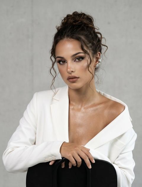 Сохранить внешность на 100%. Фото в стиле Vogue, с акцентом на чёткость и элегантность, атмосфера высокой моды. Девушка сидит на чёрном стуле (развёрнутом наоборот), поза расслабленная, уверенная, опора на обе руки. Взгляд прямой, добавляет глубины. Камера настроена на резкость, передаёт каждую деталь. На ней белый оверсайз-костюм (пиджак и брюки) классического кроя; пиджак сидит по фигуре, одно плечо открыто, без рубашки — образ дерзкий и современный. Кожа с лёгким загаром. Волосы собраны в небрежный пучок, у лица завитый локон. Макияж нюдовый: тонкие чёрные стрелки, пышные ресницы, матовые нежно-коричневые губы, хайлайтер, сияющая кожа; на декольте шиммер. Высокая детализация текстур. На ухе объёмная серебряная серьга-манжета. Освещение мягкое, направленное, с акцентом на лицо и украшения, контраст с минималистичным светлым фоном.