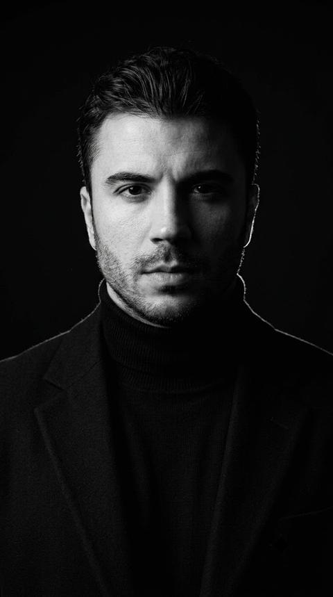 A photo of a man in a studio, standing directly in front of the camera, looking directly into the lens. The shot is from the top of the head to the middle of the chest, with the camera positioned at eye level. The main focus is the face, especially the illuminated half of the face and the eyes; the right side of the face gradually fades into deep shadow.

The natural, detailed texture of the skin is noticeable on the face.

The man is wearing a black turtleneck made of thick matte knit and a black classic jacket made of thick matte fabric. The jacket has sharply defined lapels, structured shoulders, and neat stitching along the edges. The fabrics absorb most of the light, without a pronounced sheen.

The background is completely black, uniform, without visible texture or detail. The man is positioned in front of a dark studio backdrop, with the left side of the figure separated from the background by a subtle contour light along the shoulder and the edge of the head.

The lighting is hard, directional, and high-contrast. A single, powerful light source is positioned to the left of the camera, slightly above eye level, illuminating only the left half of the face and the front of the shoulder. The right half of the face is almost entirely in deep black shadow. The transition between light and shadow is sharp, with minimal shadow fill. The overall exposure is intentionally low, immersing most of the frame in deep black.

Shot with an 85mm portrait lens, shallow depth of field, focus point on the visible eye and face, and priority focus on the face. The background is completely blurred and visually blends into the black space. Highly detailed textures of the skin, knitwear, and jacket fabric are captured. Black and white studio photography, deep tonal scale, hard directional lighting, highly detailed, photorealistic, cinematic editorial portrait photography, monochrome.