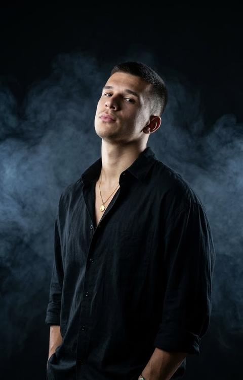 The man stands in profile, slightly turned toward the camera, his chin raised, looking into the camera. The shot is from mid-thigh to the crown of his head.

The man is wearing a loose, voluminous, oversized black shirt made of lightweight cotton fabric with a matte finish (RGB #F5F5F2), the top buttons undone, and the sleeves rolled up to the elbows. Around his neck is a thin gold chain with a small round pendant with a smooth, glossy surface (RGB #D4AF37). On his wrists are several thin fabric bracelets of various colors with a matte texture. His loose trousers are made of thick cotton with a matte finish.

His hands are tucked into his pockets—a pose of casual elegance and quiet confidence.

The background is deep, inky black, and the scene is filled with misty, bluish-gray smoke that swirls around the man, creating an atmosphere of mystery and drama.

The lighting is focused and dramatic, with a spotlight illuminating the man's face and upper body. The image has a cinematic quality. The color palette is dominated by dark, muted tones, creating a striking contrast with the man's expressive features. Style: Dark Academy, Noir, Dark, Cinematic.