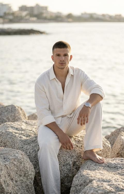 A man sits on large coastal boulders by the sea. One leg is bent and rests on a rock in front of him, the other is extended down between the rocks. One hand rests on his knee, the other hangs loosely by his side. His body is slightly turned to the side, his gaze directed toward the camera. The shot is taken full-length, at eye level, with the composition centered on the man's figure amid light-colored rocks against a backdrop of calm water and the horizon.

The man is wearing an oversized soft white (#F5F4EF) linen shirt made of fine linen with a matte textured surface. The shirt has long sleeves, slightly rolled up to the forearms, an open collar, and a loose fit. The wide-leg trousers are made of soft white (#F4F3EE) linen with a matte textured surface. The fabric folds naturally and drapes softly over the figure. On his wrist is a watch with a silver-tone metal case (#C8CCD2) and a smooth strap. Shoes are missing.

The scene takes place on a rocky seashore. Large, light-gray granite boulders (#B8B2A8) with a rough, natural texture surround the model. In the background is a calm water surface with soft reflections of sunlight. In the distance, a low-lying shoreline with silhouettes of buildings is visible. The space appears open, calm, and filled with the atmosphere of a warm seaside evening.

The lighting is natural, warm, and directional. The main light source is positioned low above the horizon, to the side of the model. The sunlight softly illuminates the face, hair, and clothing, creating a smooth volume and highlighting the texture of the linen fabric and stones. The shadows are soft and elongated. The contrast is moderate. The light retains detail in both the highlights and shadows.

Shot with an 85mm portrait lens. Shallow depth of field. The focus is on the man's face and upper body. The foreground rocks and figure remain sharp, while the distant shoreline and horizon gradually fade into a soft blur. Highly detailed linen fabric, stone surfaces, water, and natural light are captured. Coastal lifestyle portrait photography, highly detailed, photorealistic.

Warm coastal sunset mood, soft ivory and warm stone dominant tones, muted natural palette, medium-low saturation, low to medium contrast, gentle golden hour lighting, smooth shadow transitions, delicate highlight rolloff, airy seaside atmosphere, soft film-like rendering, rich linen texture detail, creamy background separation, minimal grain, refined travel editorial color grading, luxury coastal lifestyle photography look.